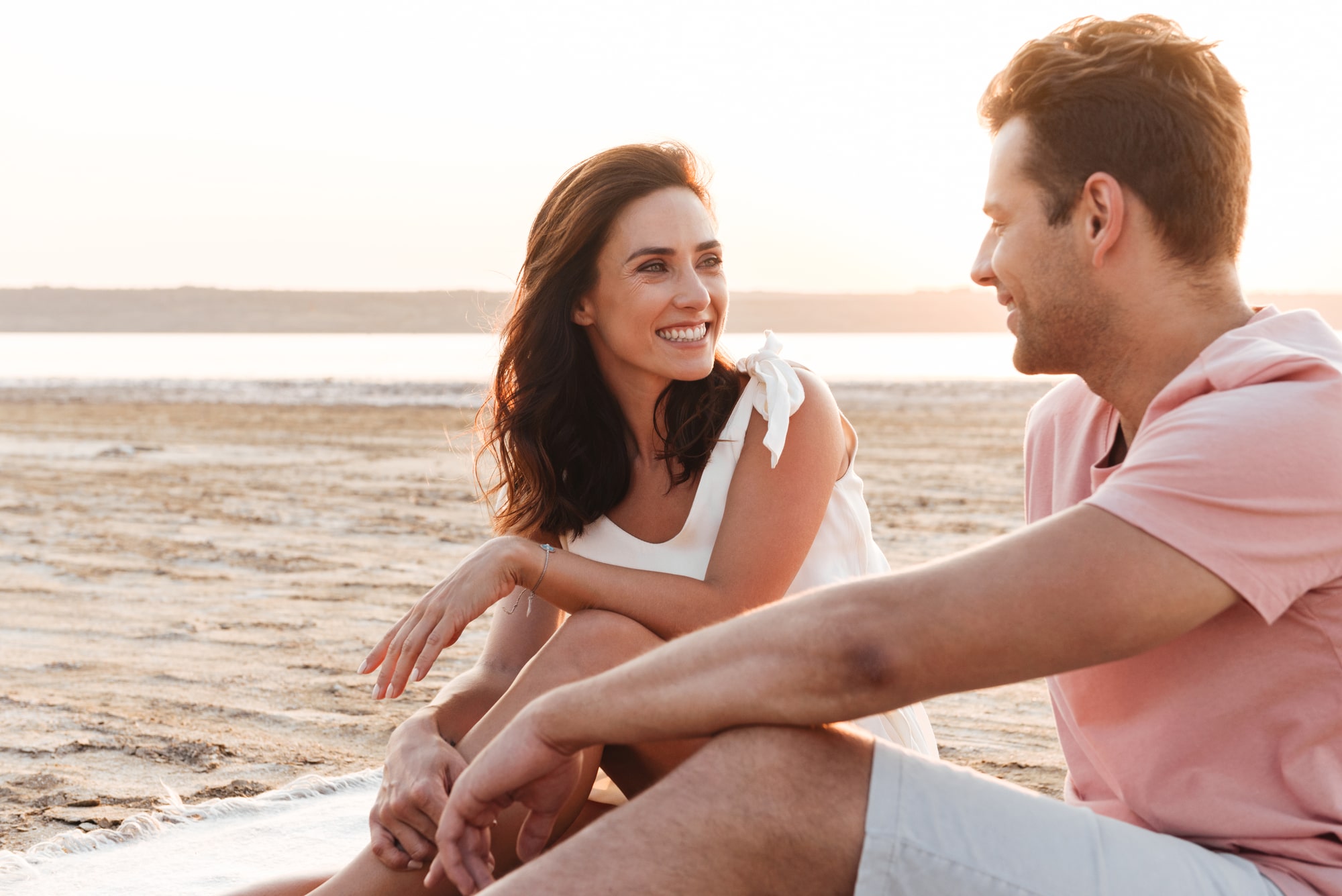 Lovely young couple wearing summer clothing relaxing on a blanket at the beach, looking at each other