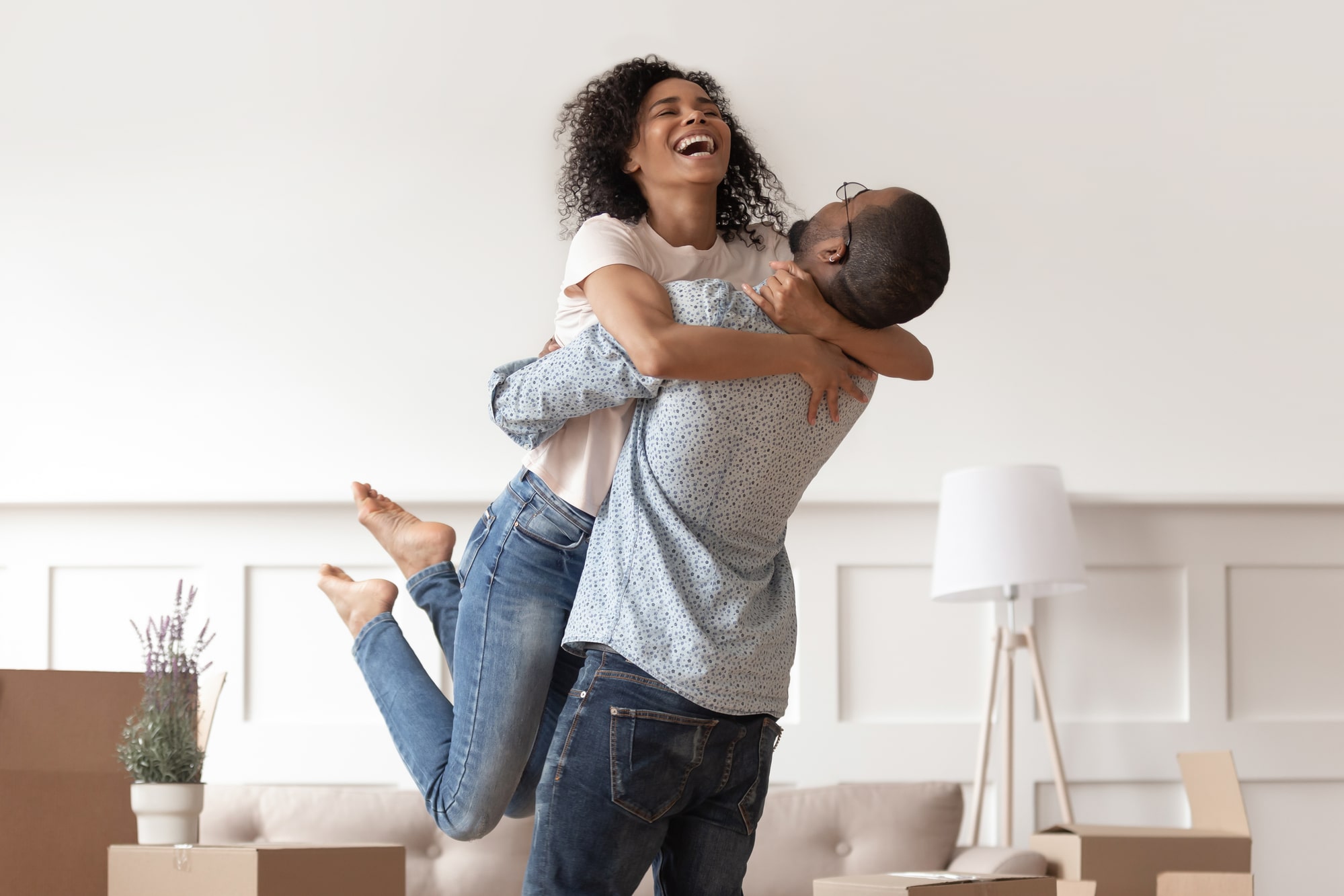 African husband lifting happy wife laughing celebrating moving day with boxes, excited first time home buyers renters owners having fun enjoy relocation into own new flat house, mortgage investment